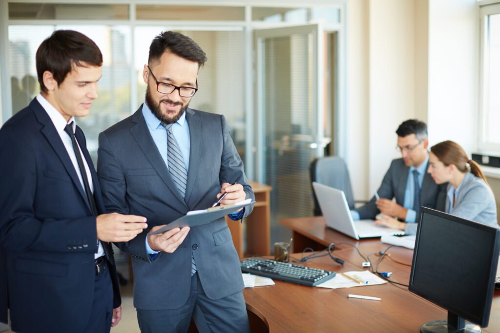 Confident businessman with clipboard consulting his partner on background of their colleagues in office
